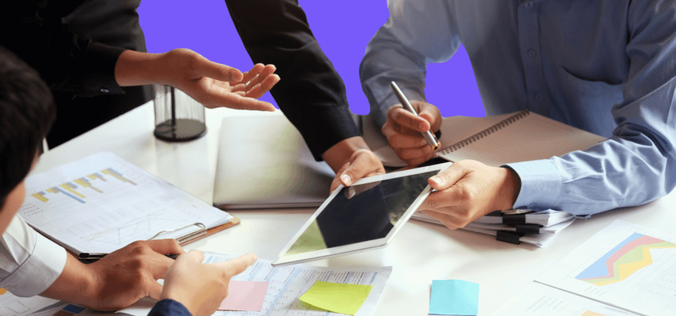 a group of people around a table with scattered papers and an ipad